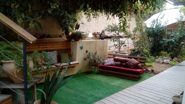 A cozy outdoor seating area with a wooden bench and a cushioned sofa, surrounded by potted plants and a small garden. - Wonderful Room Garden Apartment (Tel Aviv)