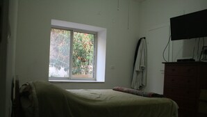 A bedroom with a single bed, a dresser, a television mounted on the wall, and a window with a view of trees. - Wonderful Room Garden Apartment (Tel Aviv)