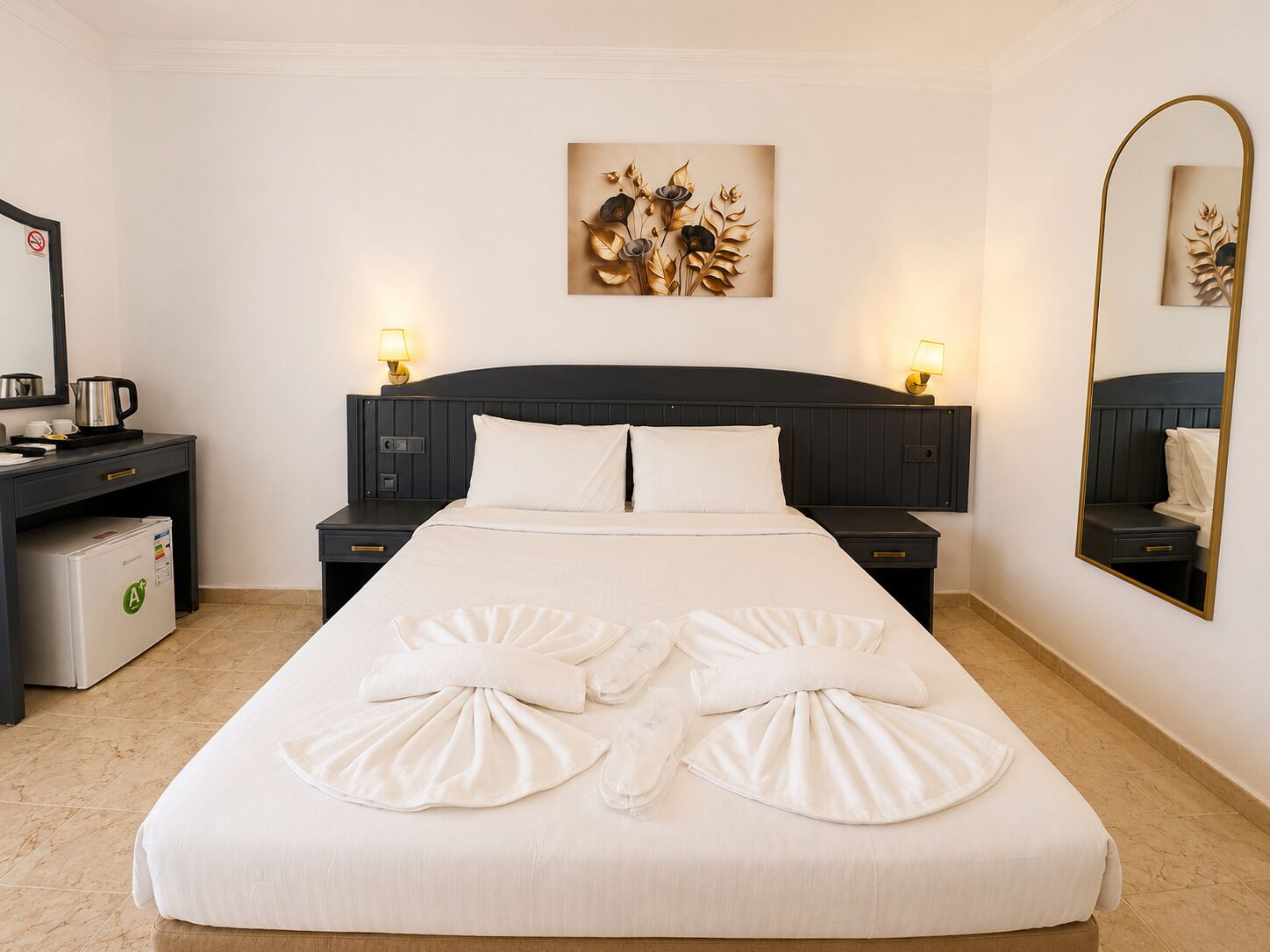 A neatly made bed with towels, a bedside table with a lamp, a small desk with a kettle, and a wall-mounted mirror.