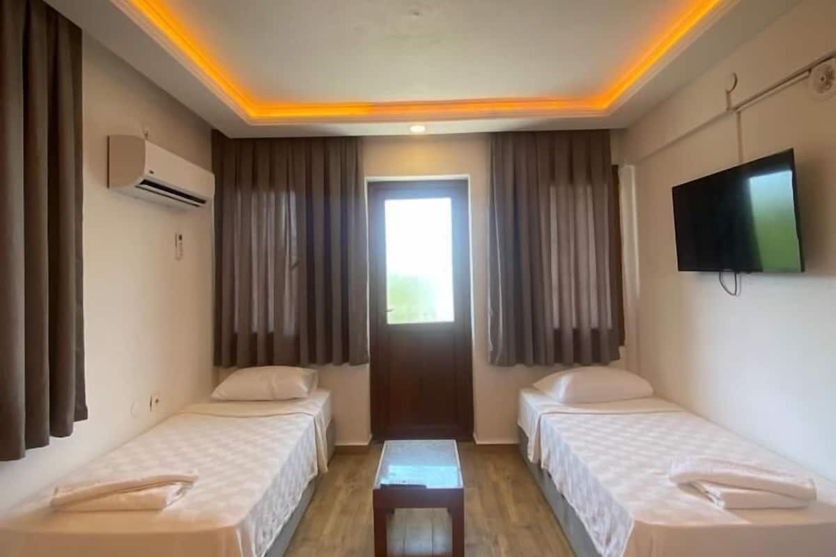 Habitación de hotel con dos camas individuales, suelo de madera, una mesa en el centro, unidad de aire acondicionado, televisor montado en la pared y una ventana con cortinas.