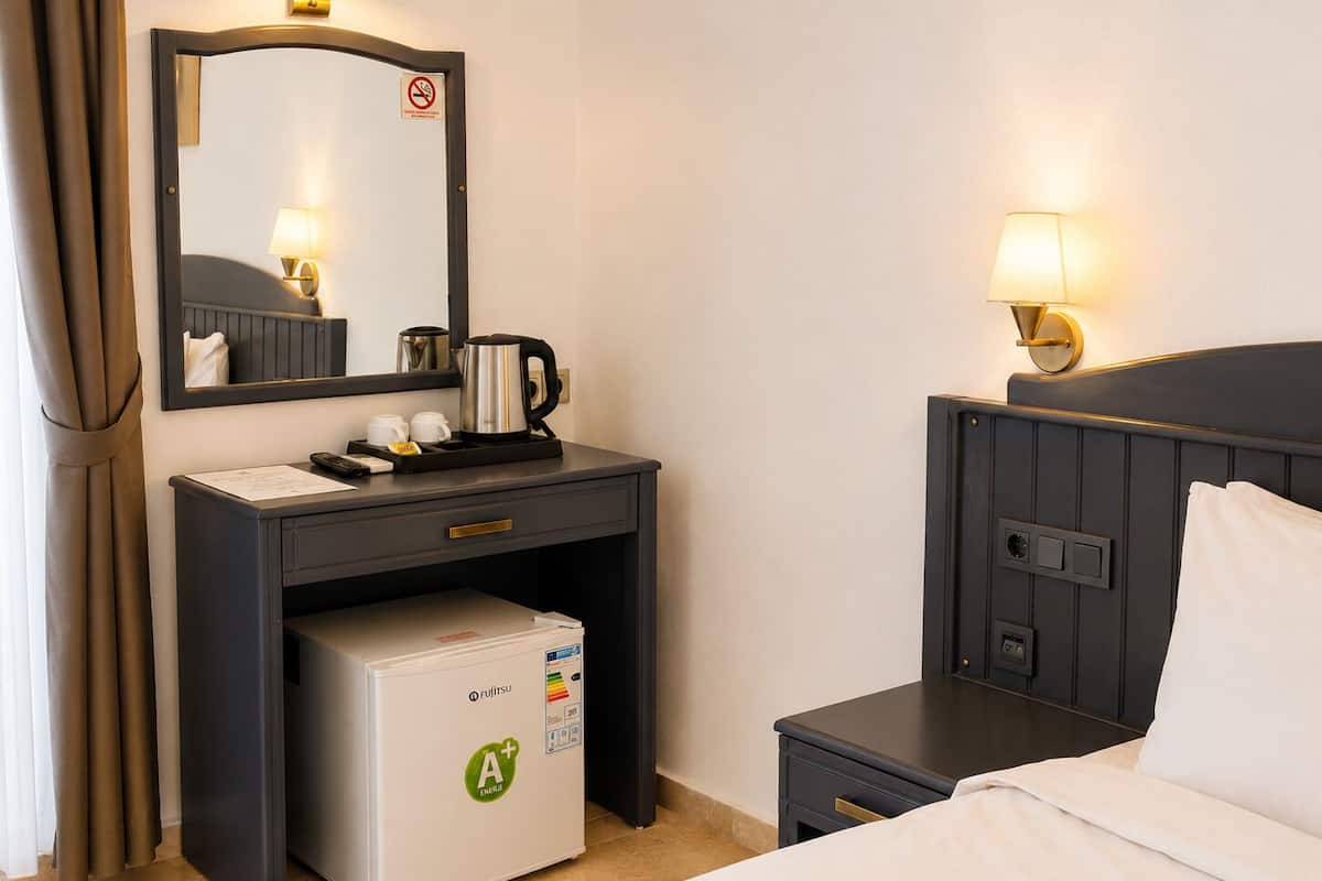 Una habitación de hotel con una cama, un escritorio con cafetera, un refrigerador, una mesita de noche y una lámpara de pared.
