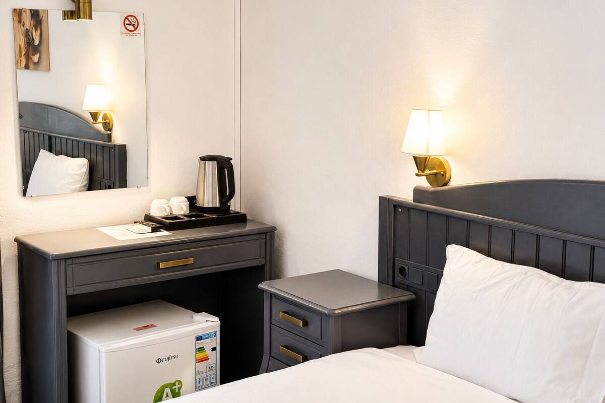 Habitación de hotel con cama, mesita de noche, un pequeño escritorio con tetera, espejo y refrigerador.