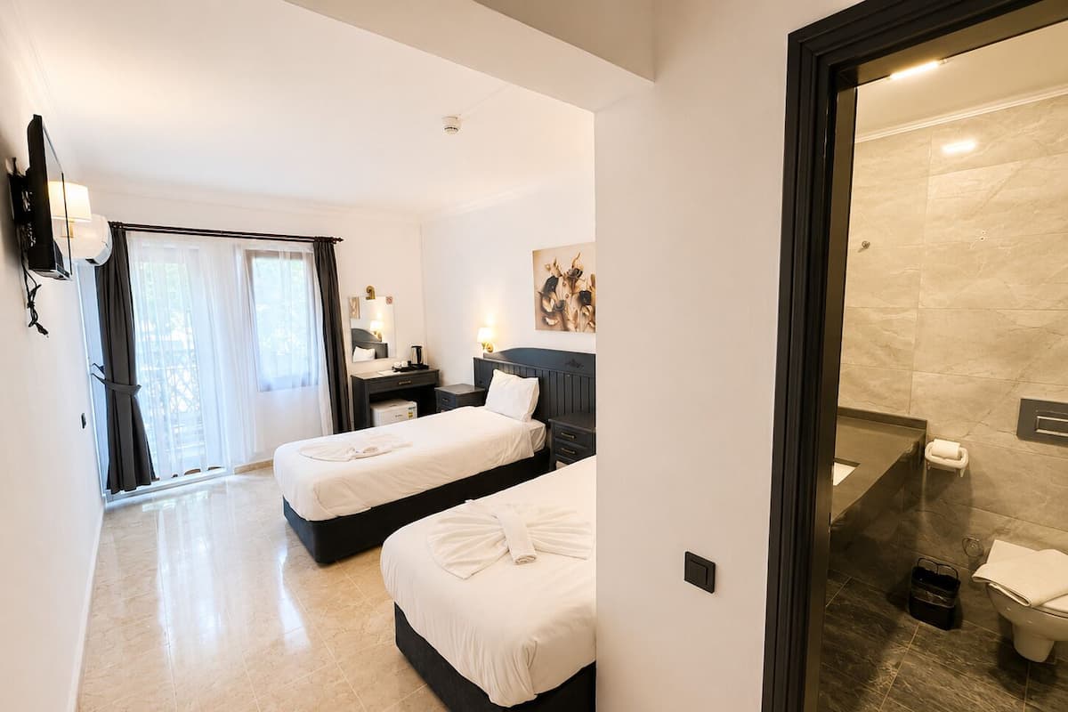 Habitación de hotel con dos camas, un escritorio, una silla, baño con ducha e inodoro, y un ventanal con cortinas.