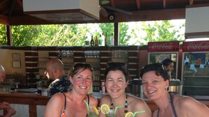 Un groupe de personnes est assis au bar, des boissons sont posées devant elles. On aperçoit un plafond en bois et, au fond, un réfrigérateur Coca-Cola.