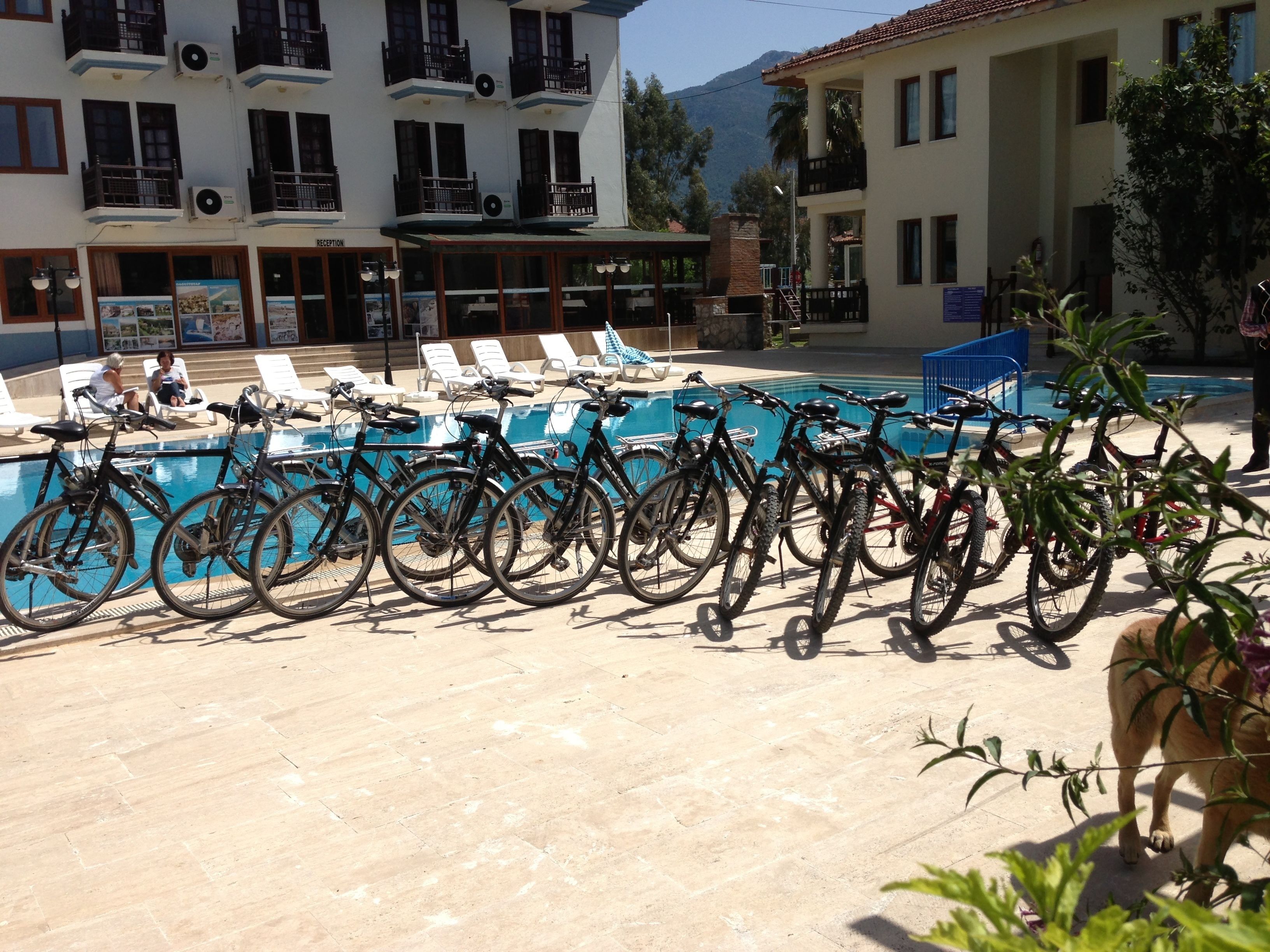 Zona de piscina con bicicletas estacionadas al frente, sillas de descanso y un edificio con varios pisos y balcones.