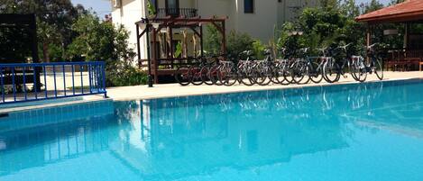 A swimming pool with bicycles parked nearby, a building with a tiled roof, and a clear sky.