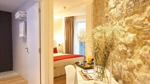 Une chambre d’hôtel avec un mur décoratif en pierre, un lit recouvert d’une couverture rouge, un coin repas agrémenté d’un vase de fleurs et une vue sur la ville par la fenêtre.