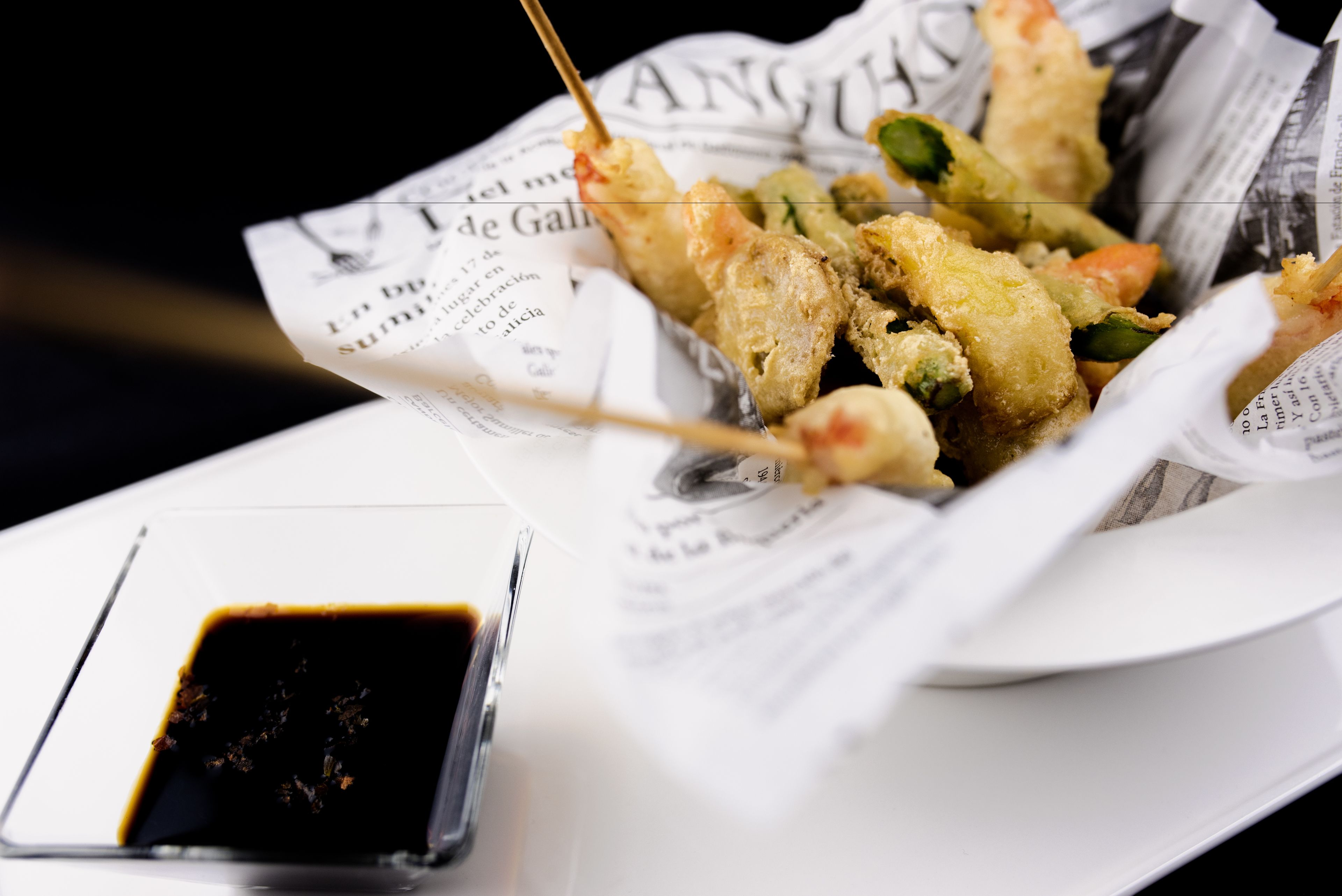 Un plato de brochetas de mariscos fritos sobre papel de periódico, con un recipiente pequeño de salsa para mojar.