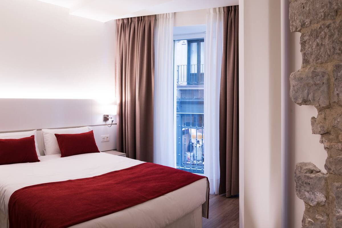 Habitación de hotel con una cama grande, almohadas y manta rojas, pared de piedra y vistas a un balcón.