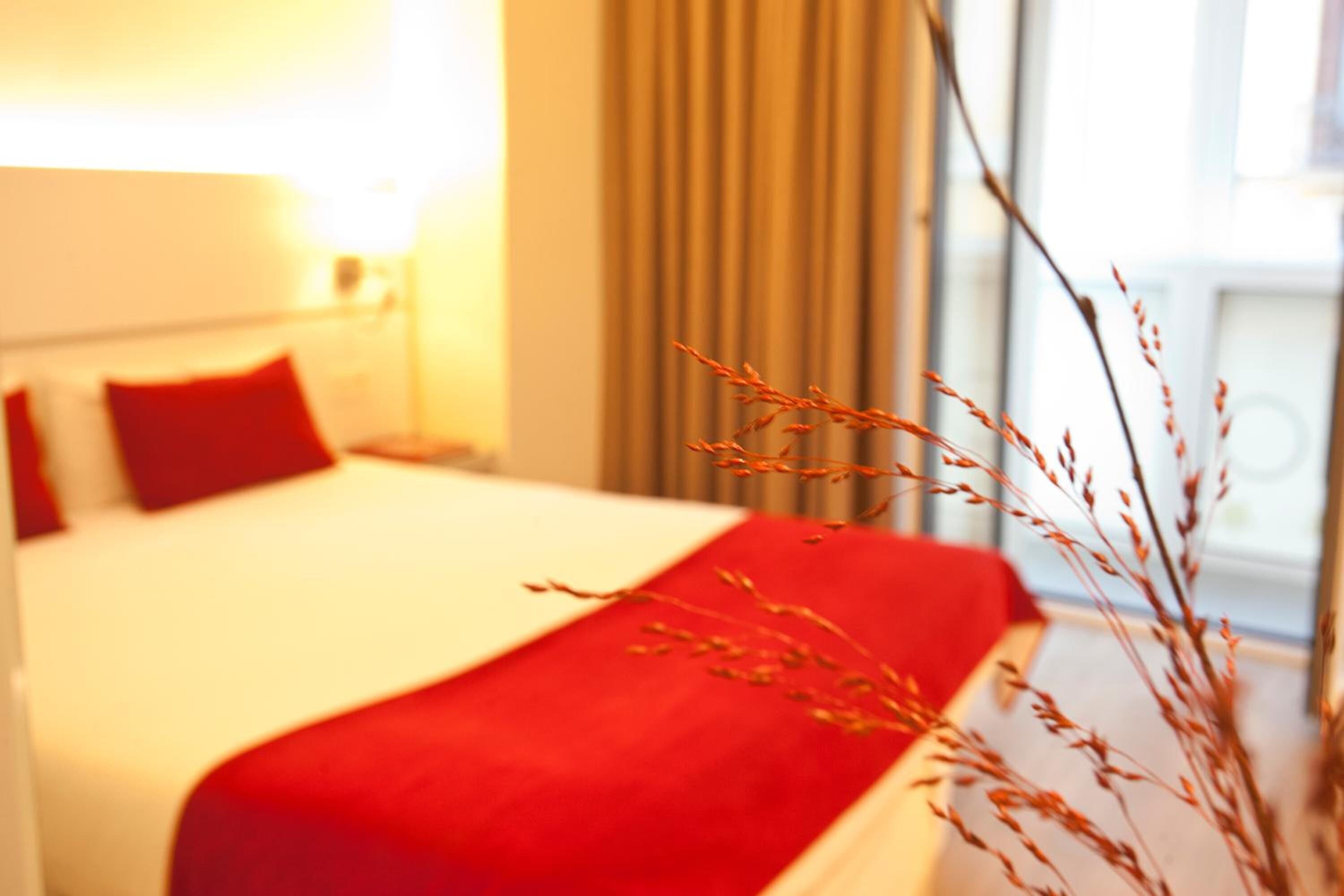 Ett hotellrum med en säng, rött sänglinne och ett fönster med gardiner.