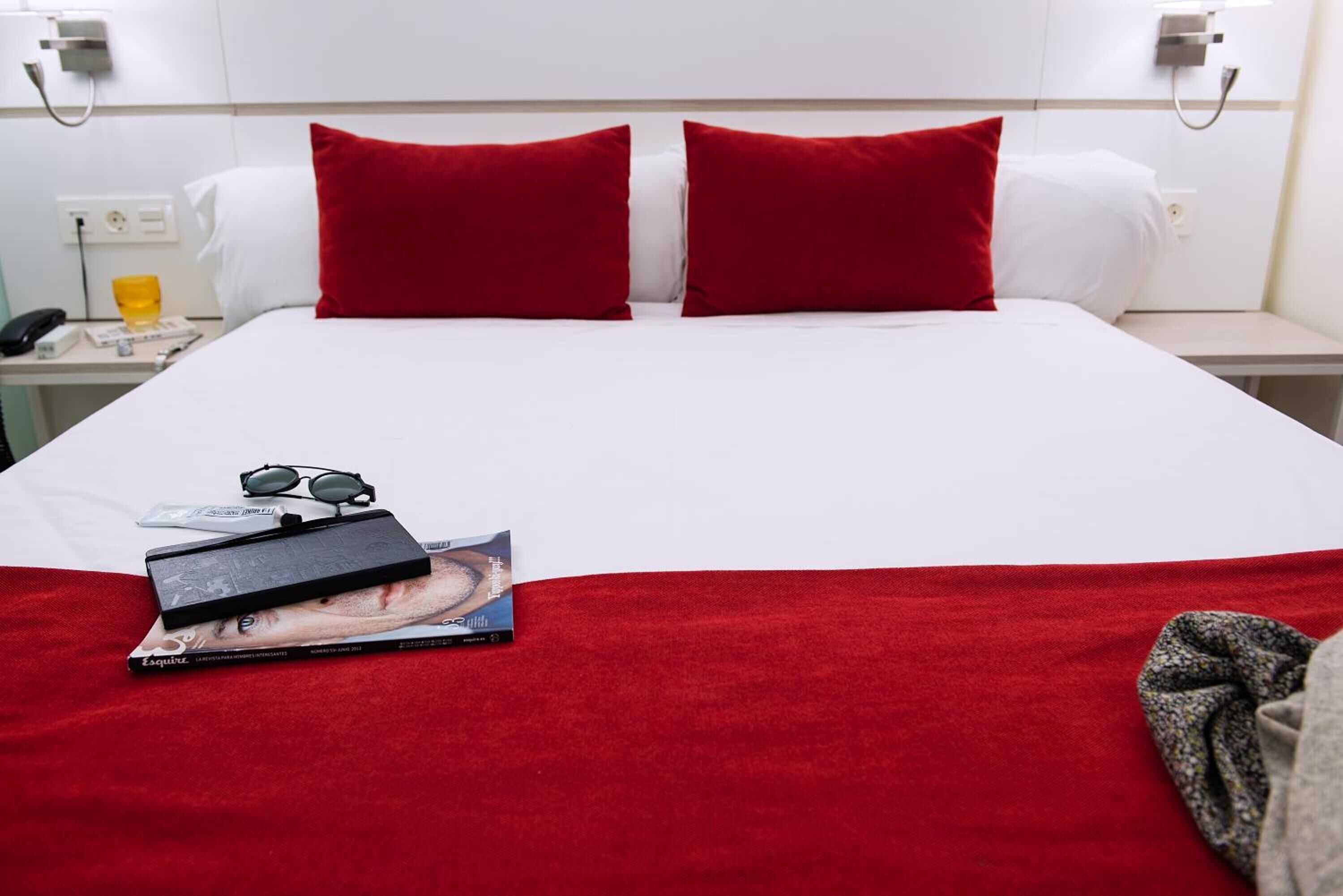 A hotel room with a neatly made bed, red pillows, a bedside table with a phone and a glass of water, and a magazine on the bed.