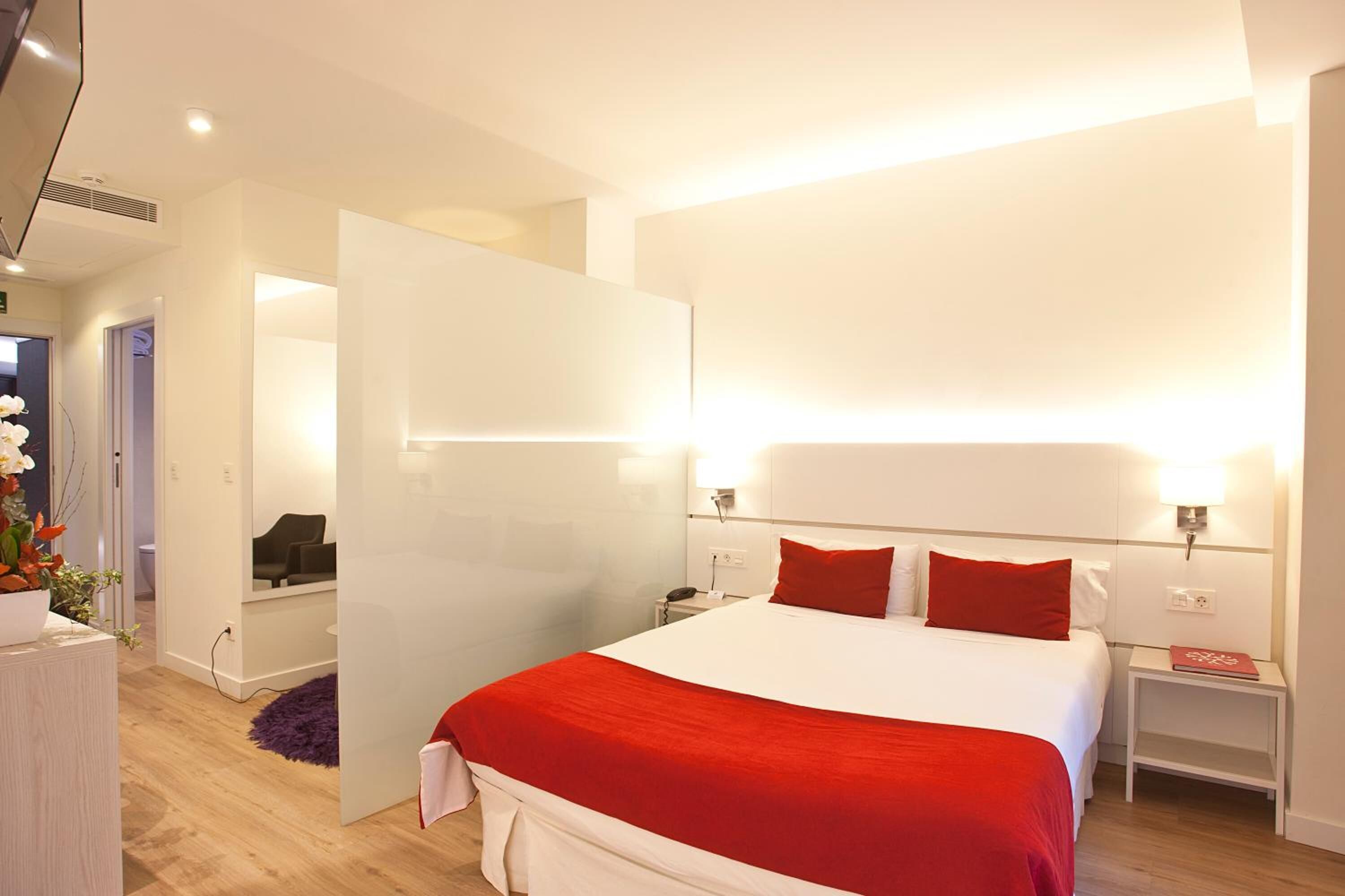 Una habitación de hotel moderna con una cama grande, paredes blancas y un detalle en rojo.