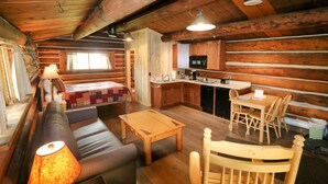 Interior acollidor d'una cabana amb taula de dinar de fusta, un sofà, una cuina petita i un llit amb llençol a quadres.