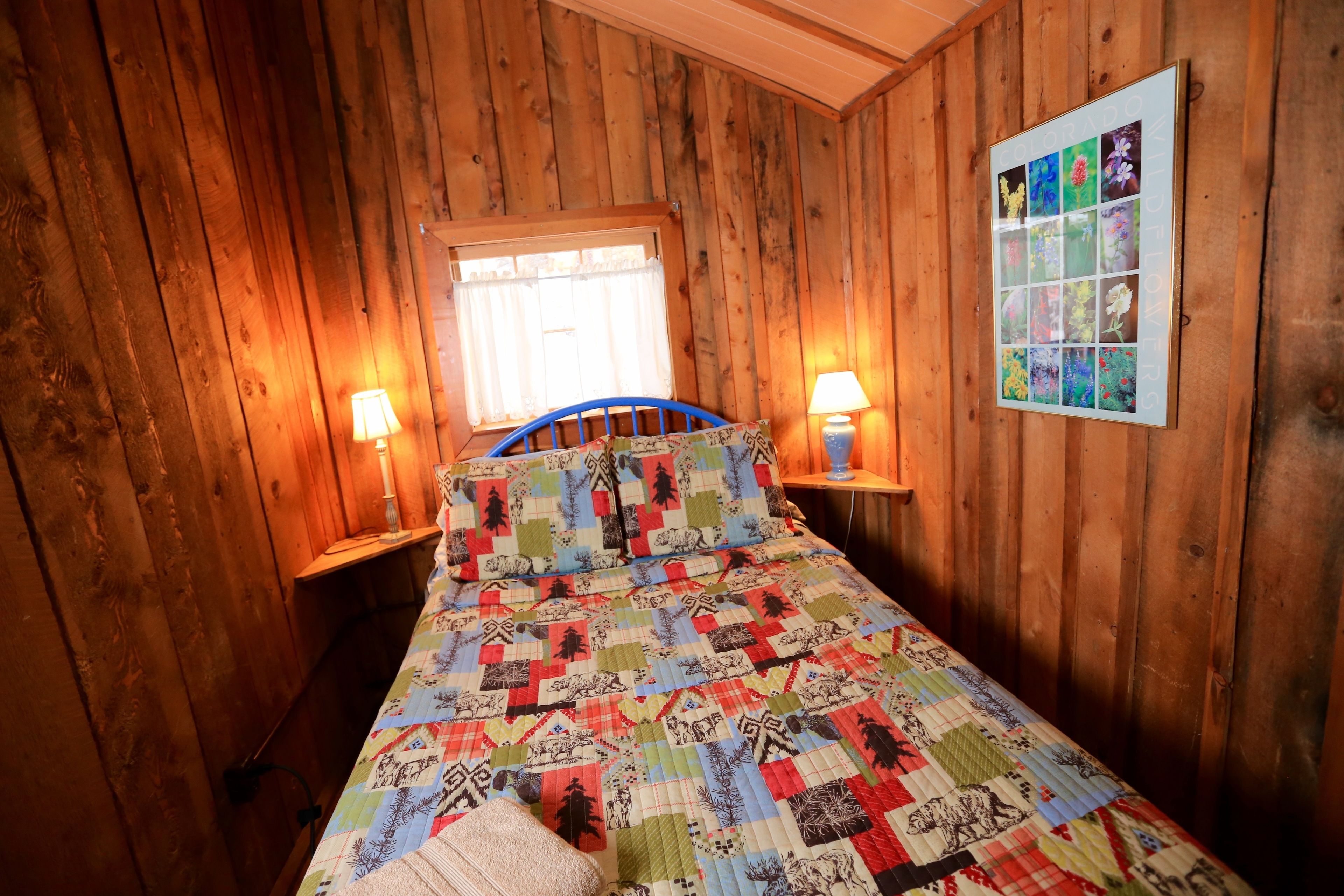 A wooden-paneled room with a bed featuring a colorful quilt, two bedside lamps, and a framed collage on the wall.