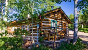 Una cabana de fusta rústica amb un porxo, envoltada d'arbres i vegetació.