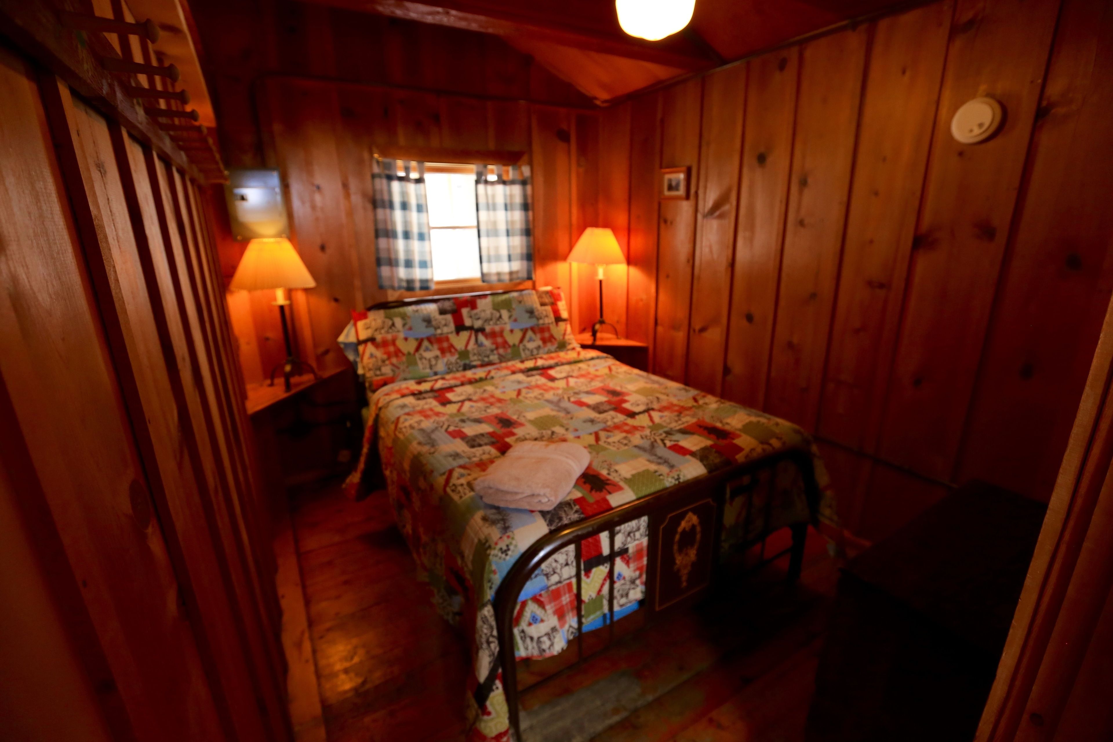 A cozy room with a wooden bed, two bedside lamps, a window with curtains, and a wooden wall.