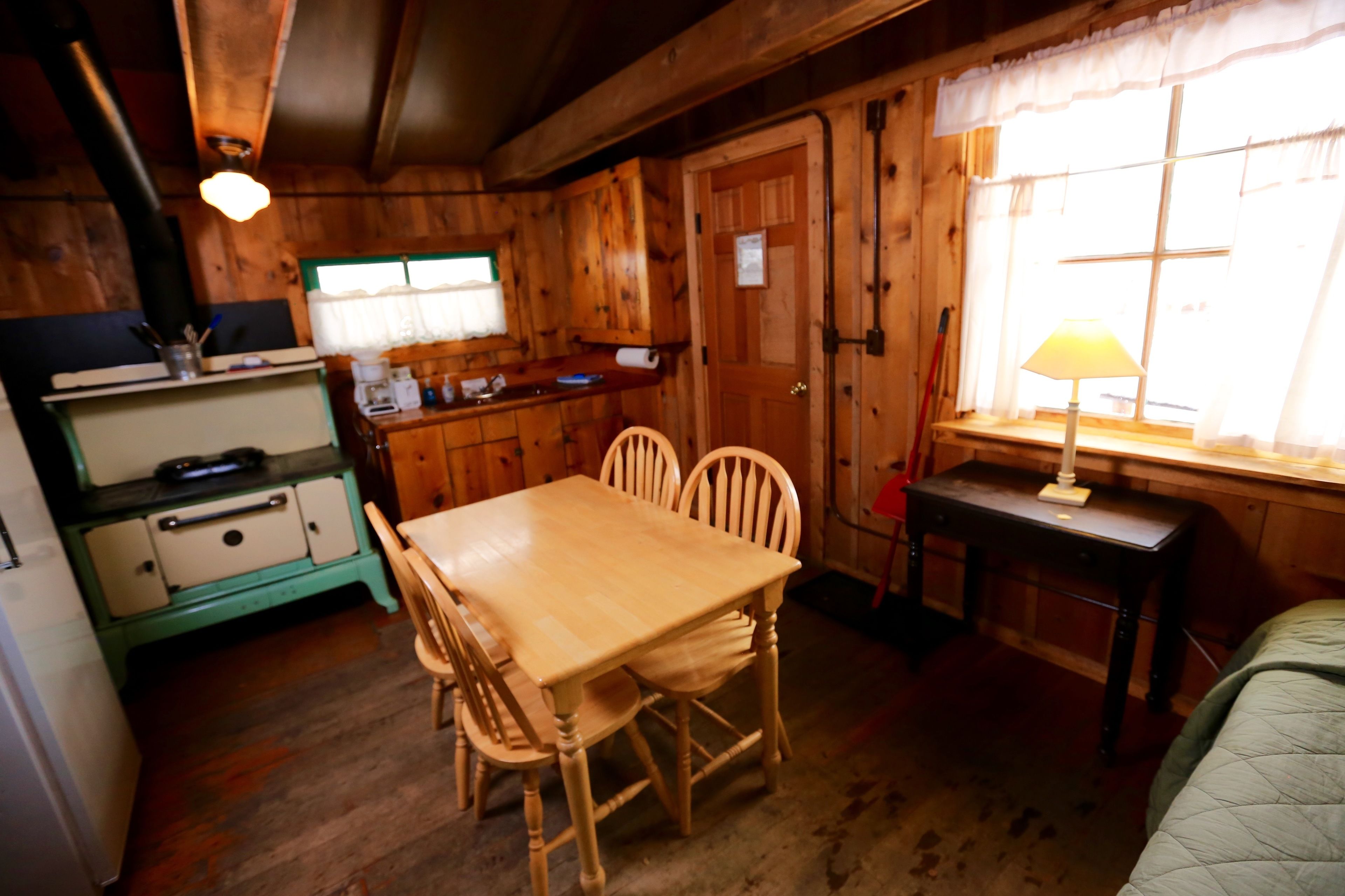 A wooden cabin interior with a dining table, chairs, a small kitchen area, and a window with a lamp.