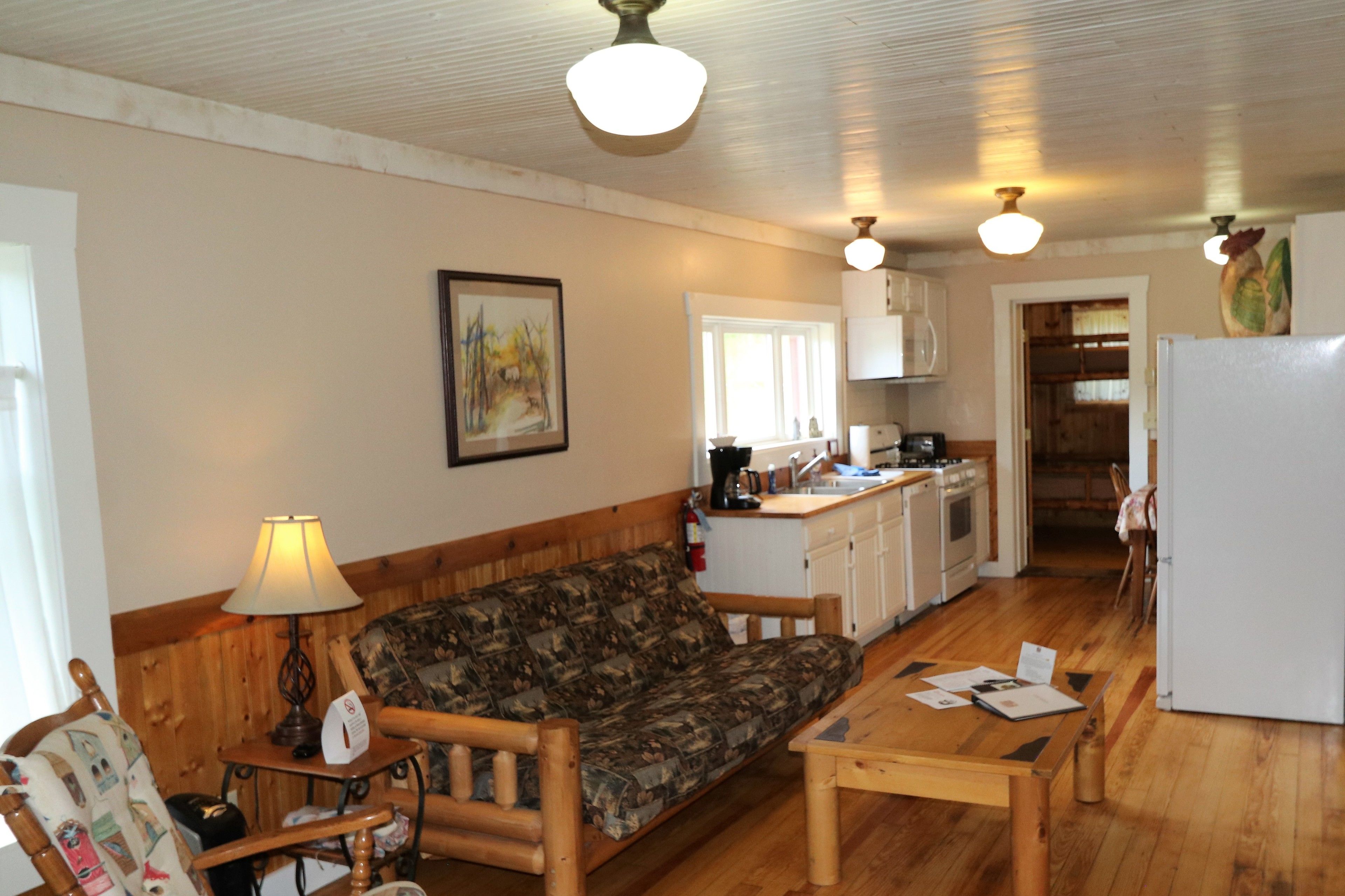 A cozy living room with a wooden sofa, a small table, and a lamp. There is a kitchen area with cabinets and a refrigerator in the background.