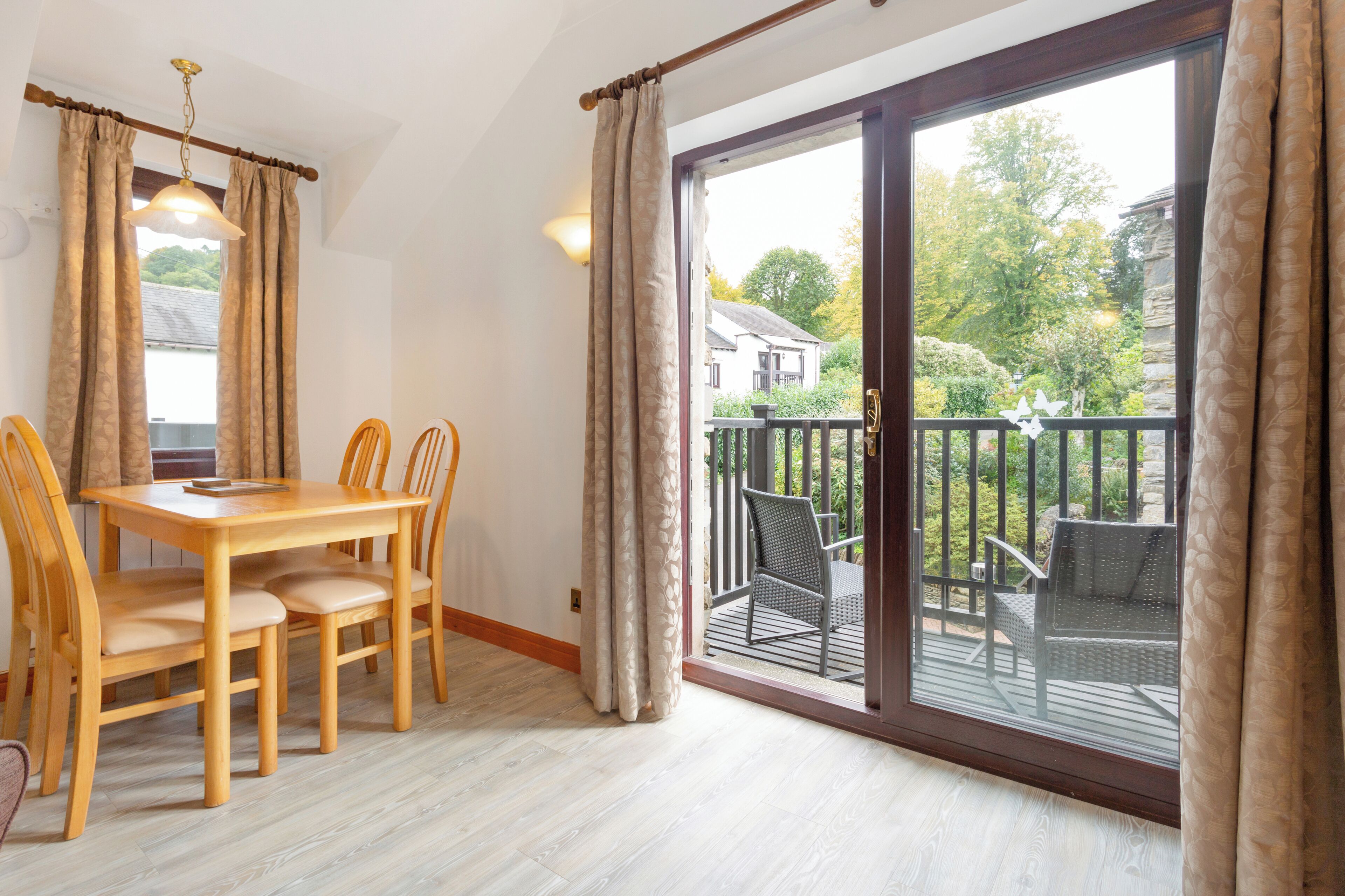 Un coin repas avec une table et des chaises, une porte-fenêtre coulissante donnant sur un balcon extérieur équipé de chaises, et une vue sur une maison et la verdure.