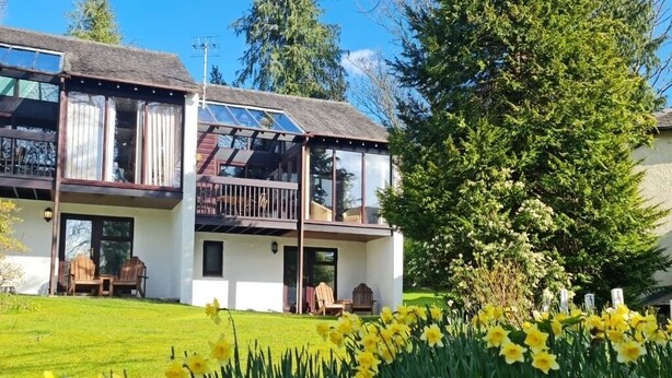 Uma casa de dois andares com uma varanda, um gramado com narcisos e uma grande árvore ao fundo.