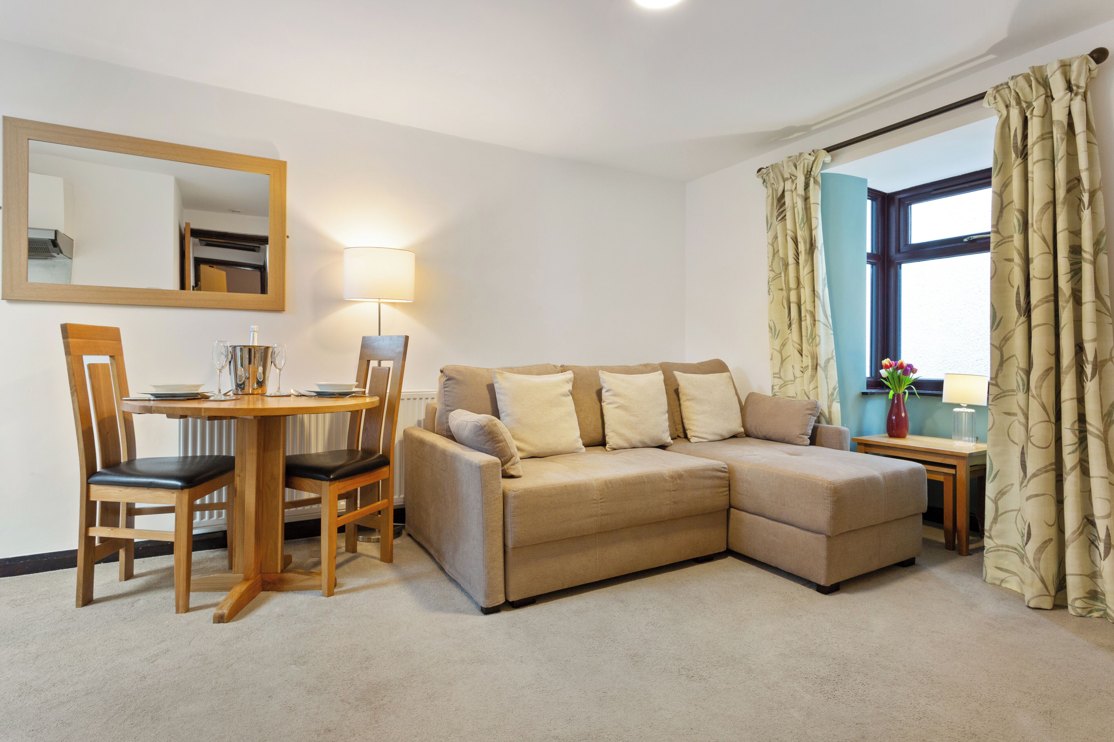 A living room with a beige sectional sofa, wooden chairs, a small table with a lamp, and a window with curtains.
