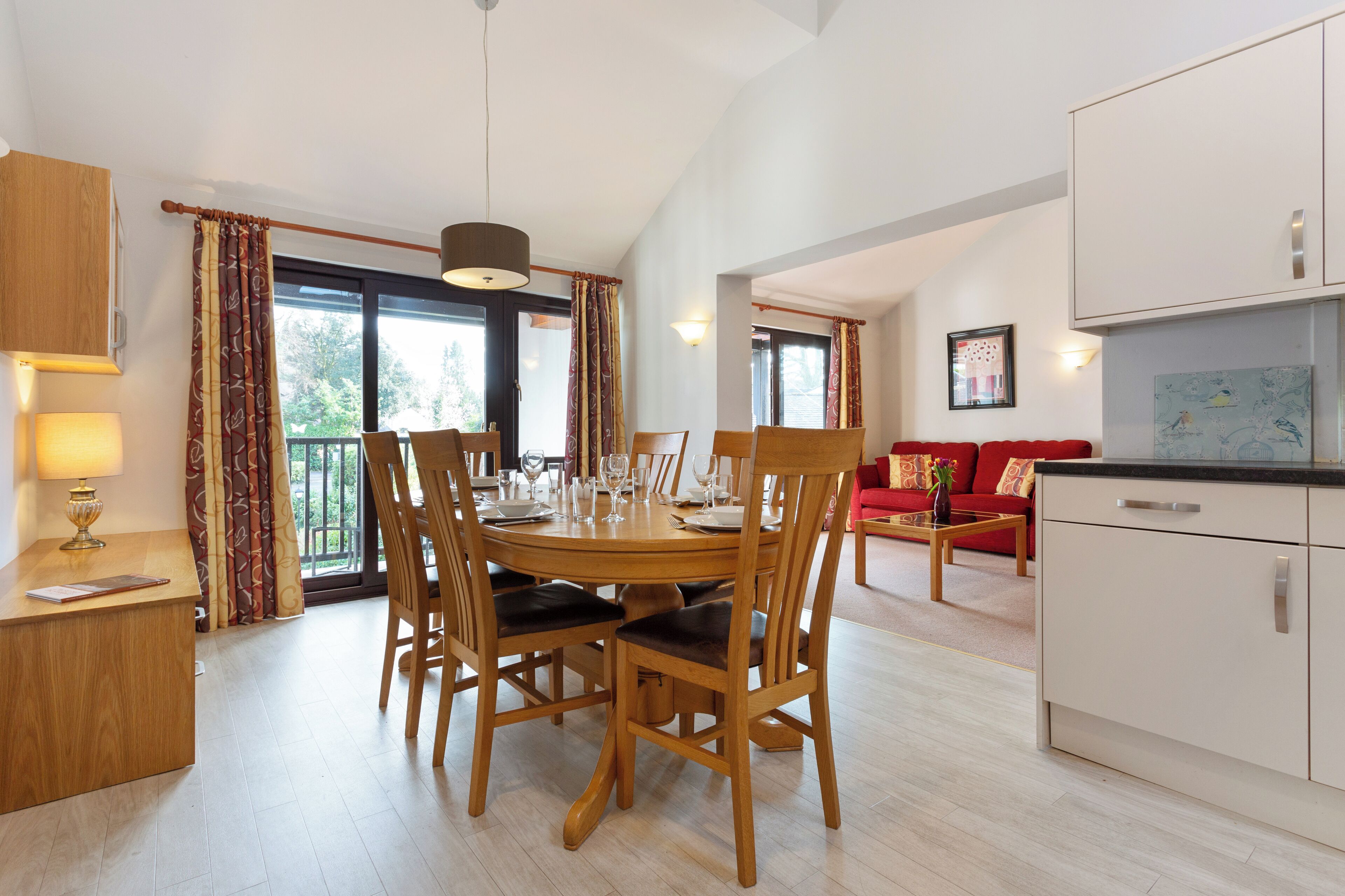 A dining area with a wooden table and chairs, a kitchen with white cabinets, and a red sofa in the background.