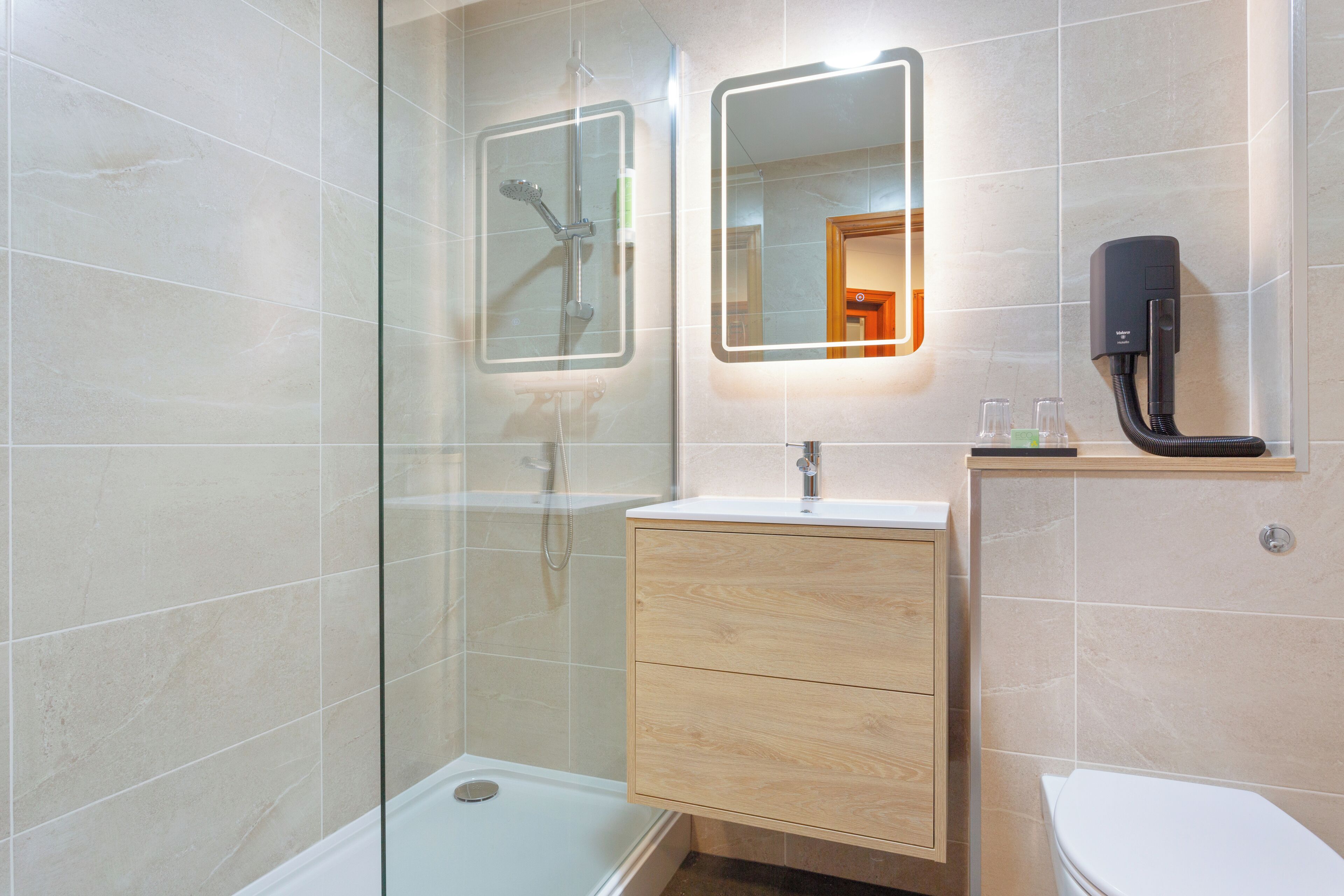 Un baño moderno con cabina de ducha de vidrio, tocador de madera, inodoro y espejo con iluminación integrada.