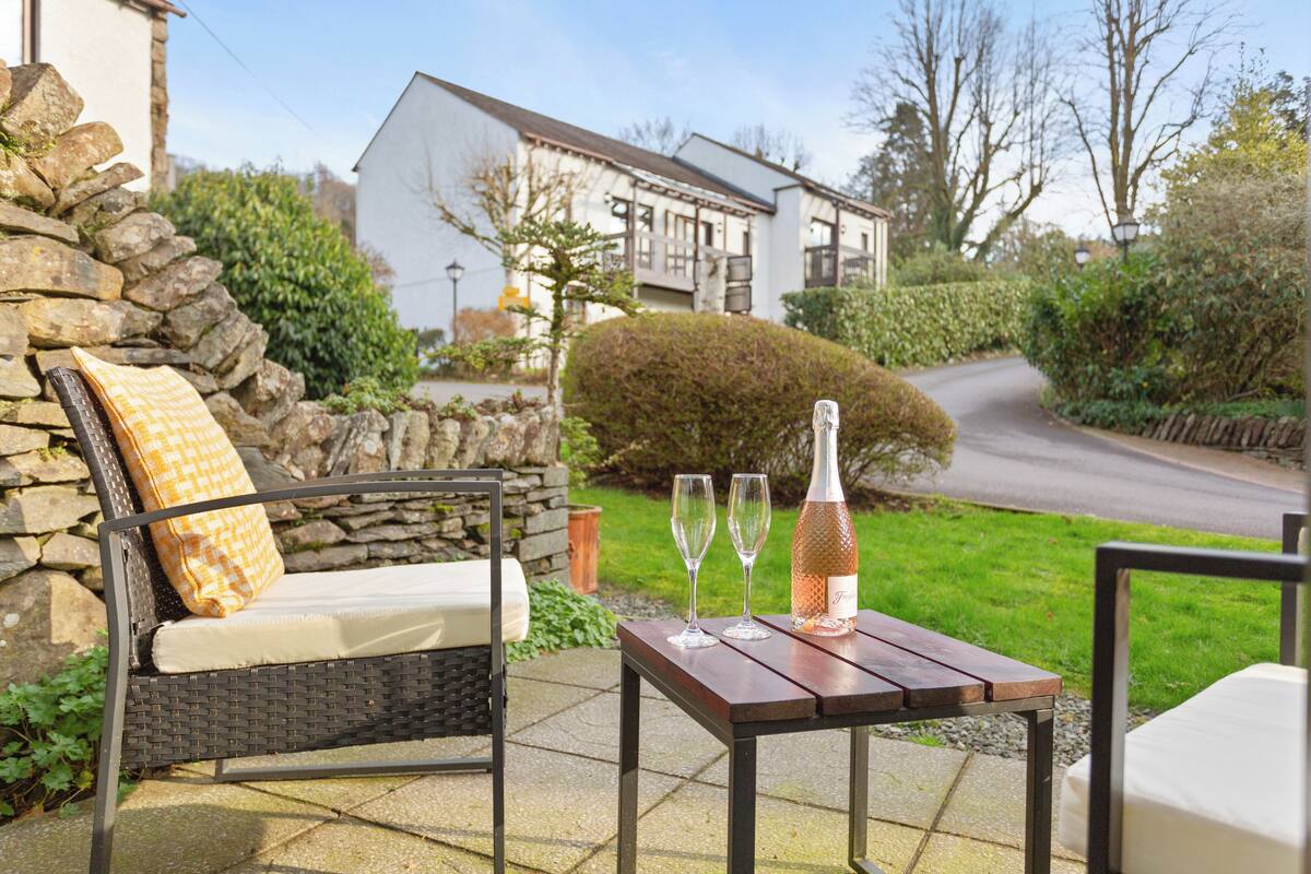 An outdoor seating area with a wicker chair, a table with a bottle and glasses, and a stone wall.