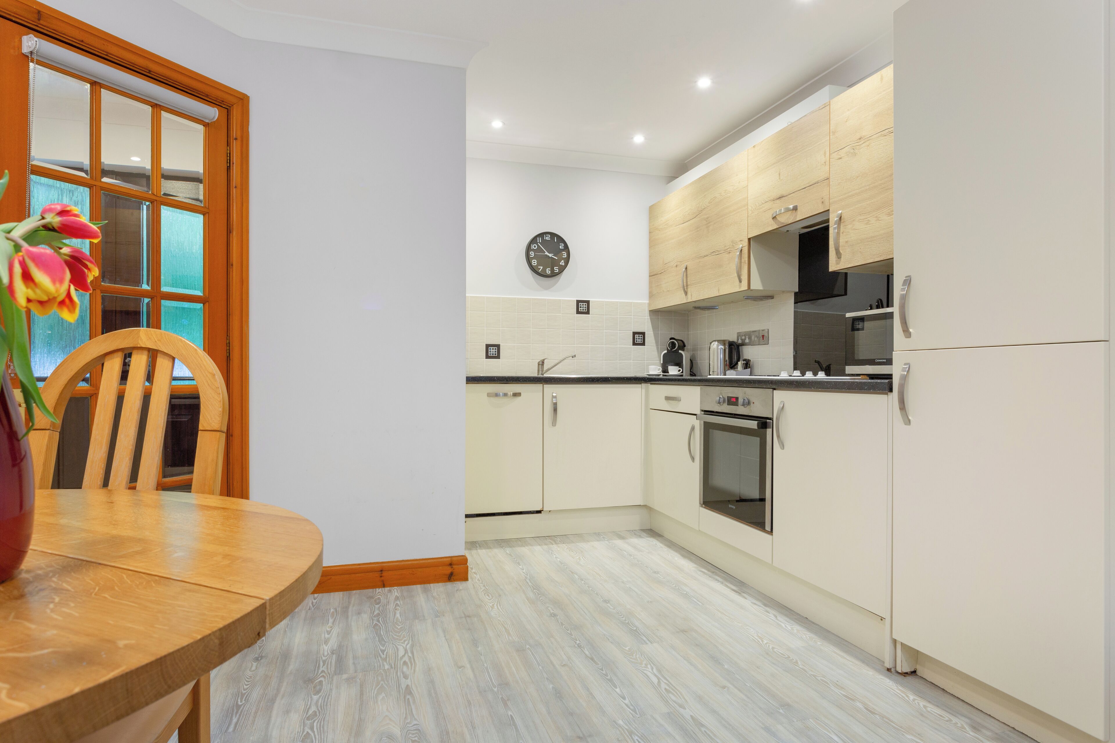 A modern kitchen with light wood cabinets, a stainless steel oven, and a wooden dining table with a vase of tulips.