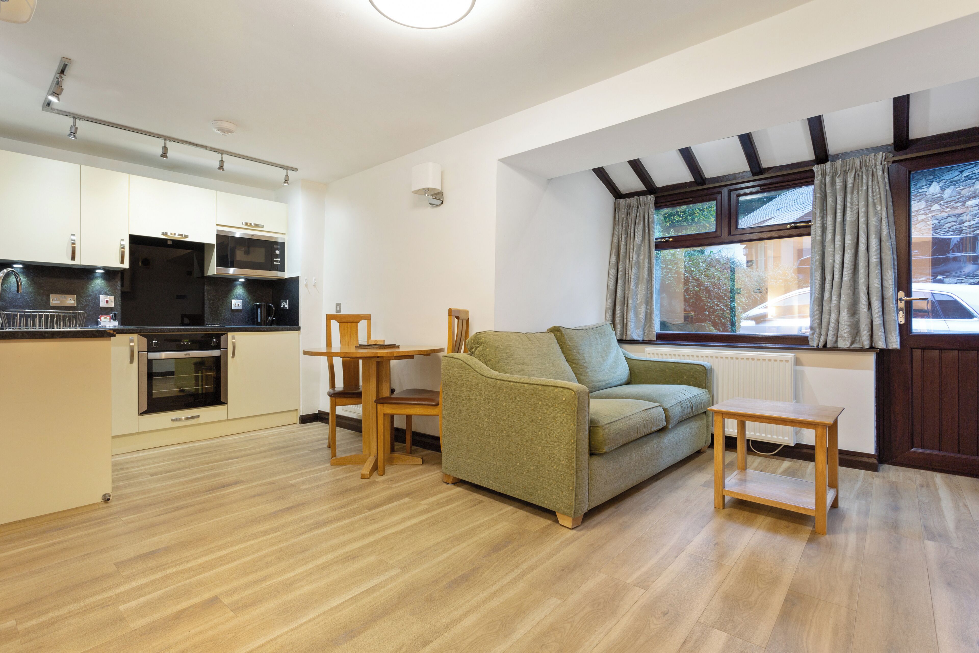 A modern kitchen with built-in appliances, a wooden dining table, a green sofa, and a window with curtains.