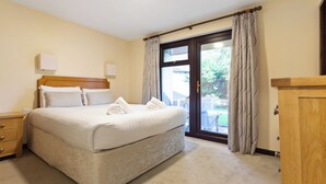 Uma cama bem arrumada com lençóis e almofadas brancas, cabeceira de madeira, mesa de cabeceira com gaveta e uma janela grande com cortinas.