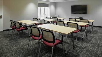 Une salle de conférence équipée de plusieurs tables et chaises, d’un grand téléviseur à écran plat fixé au mur et d’une fenêtre avec des stores.