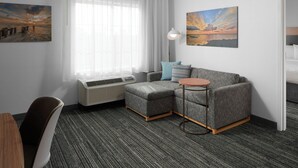 A modern hotel room with a gray sectional sofa, a small round table, a desk, and a chair. There is a large window with sheer curtains, a wall-mounted air conditioner, and two paintings.