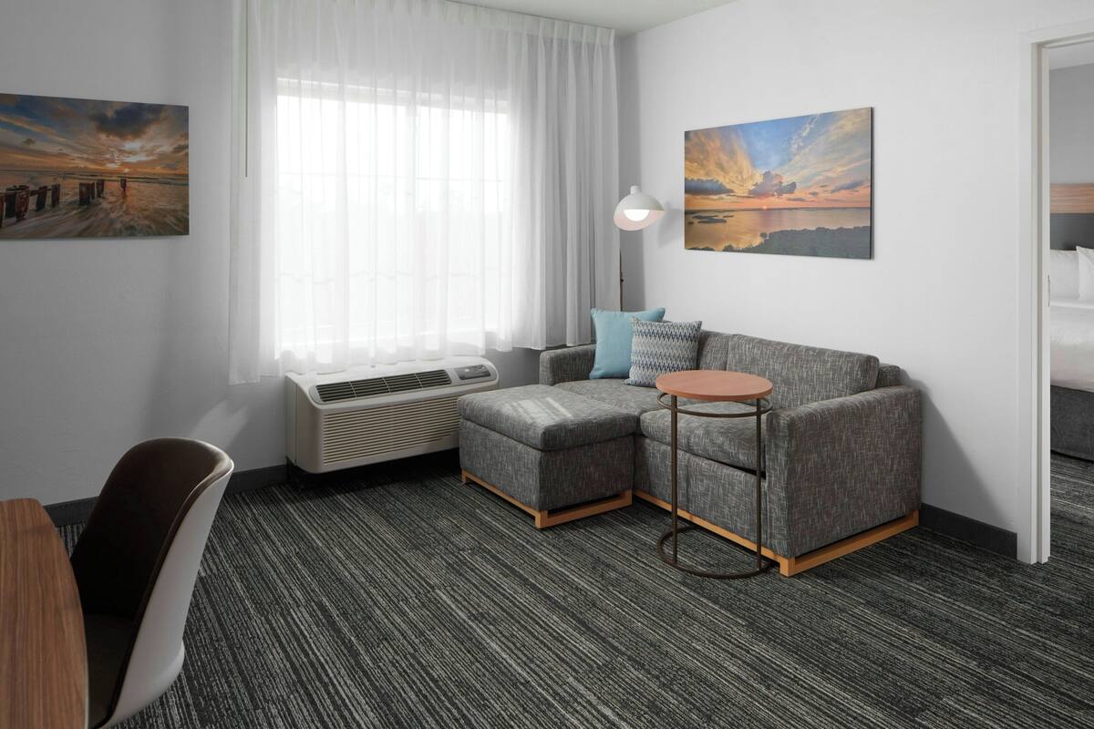 Een moderne hotelkamer met een grijze modulaire bank, een klein rond tafeltje, een bureau en een stoel. Er is een groot raam met dunne gordijnen, een wandairconditioner en twee schilderijen.