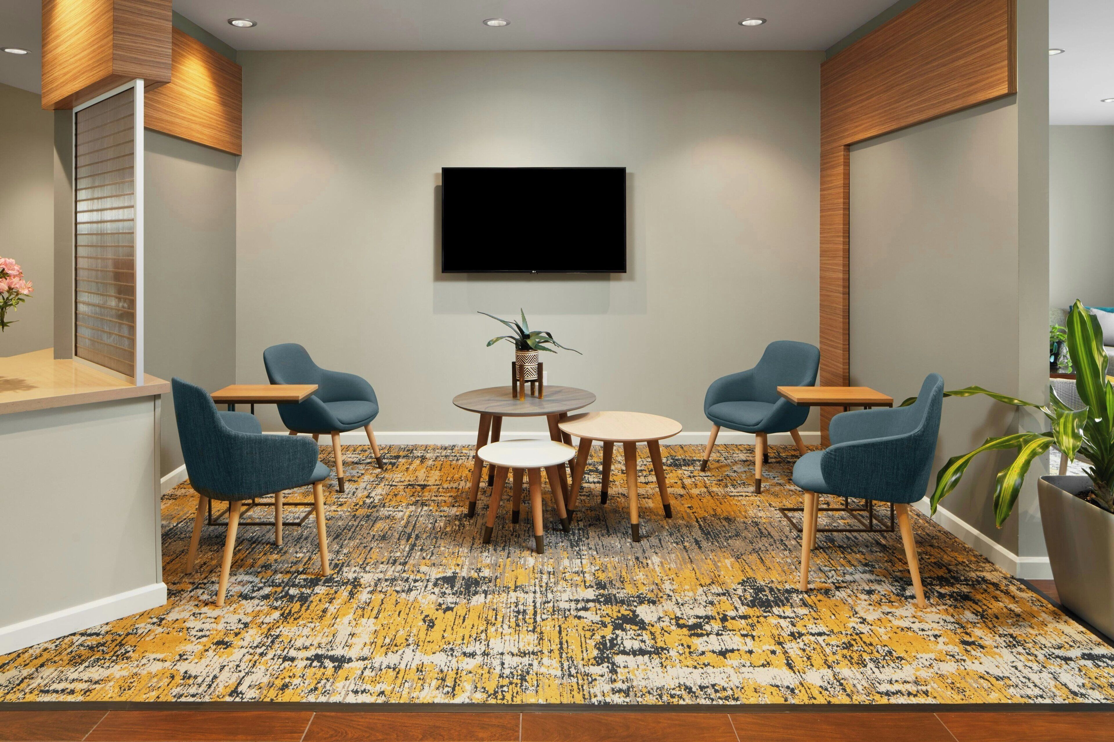 A conference room with a large flat-screen TV mounted on the wall, a round table with chairs, and a patterned carpet.