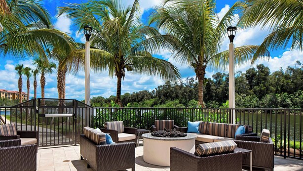 An outdoor patio area with wicker furniture, a circular fire pit, and a view of a lake with palm trees.