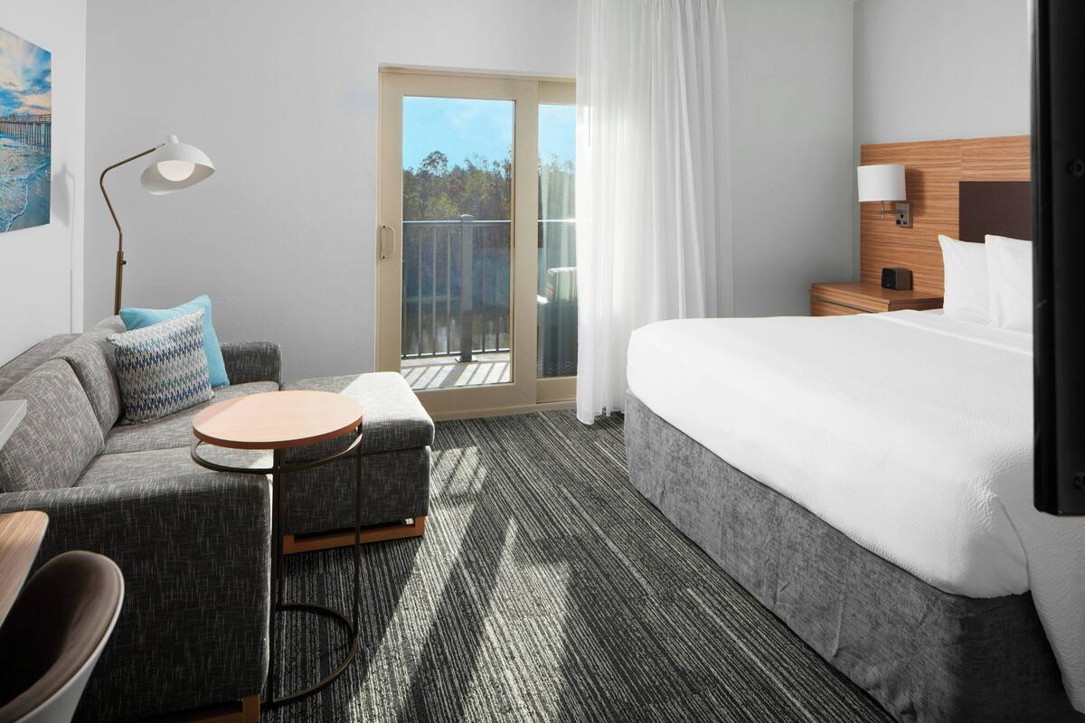 Een hotelkamer met een bed, een bank, een klein tafeltje, een lamp en uitzicht op de buitenlucht door een glazen deur.