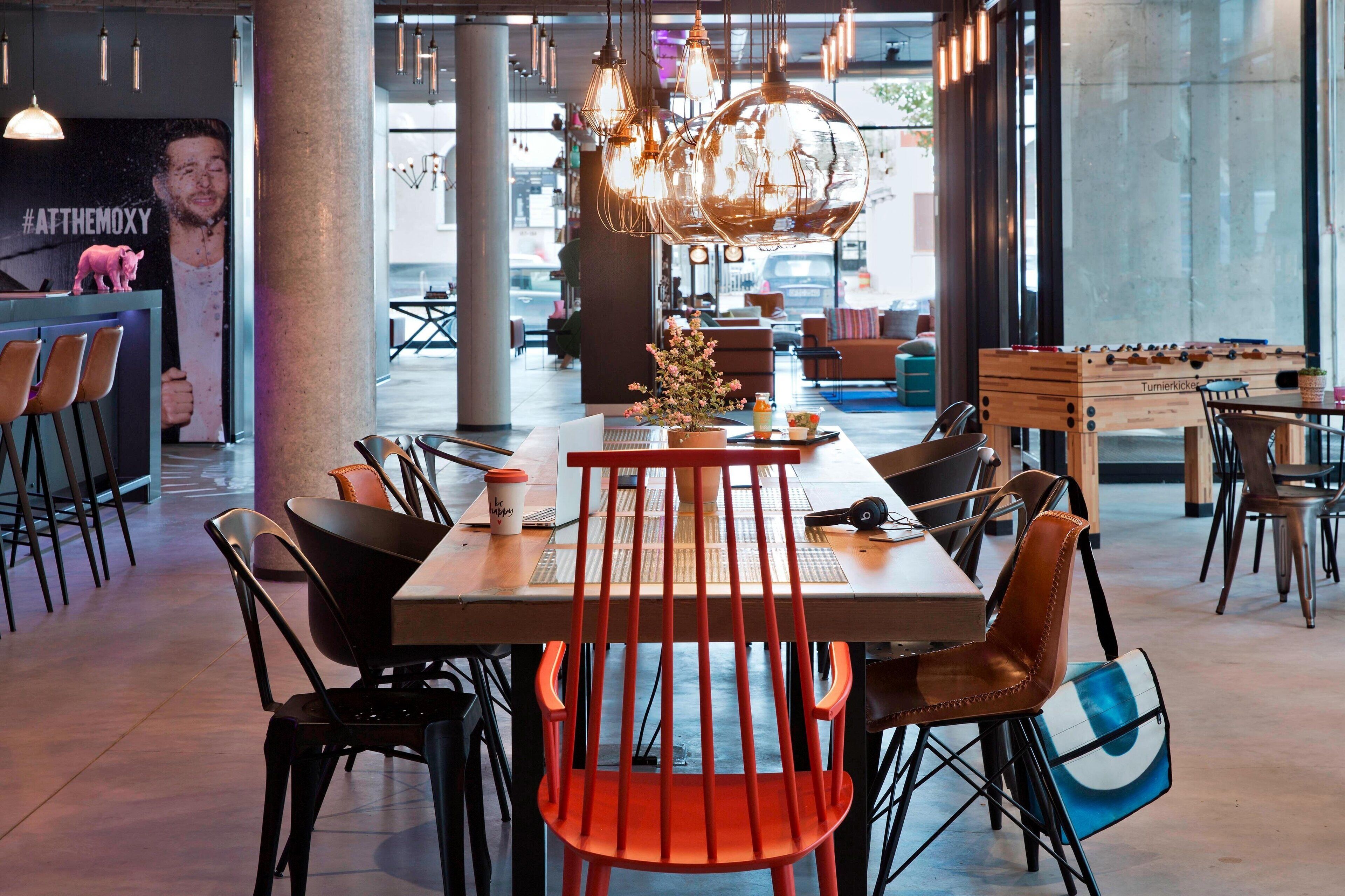 A modern dining area with a long table, orange chairs, and a foosball table.
