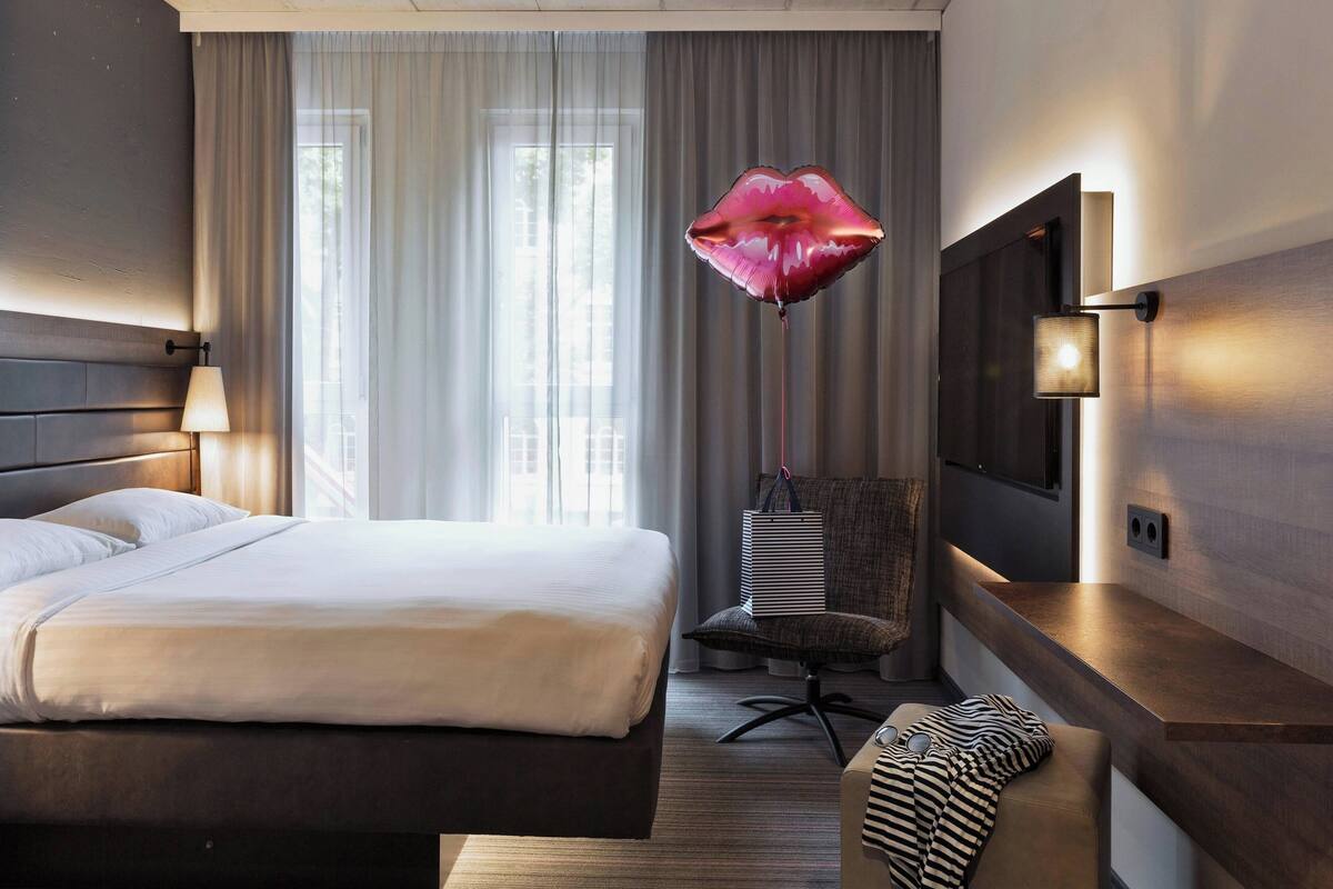 Habitación de hotel con una cama grande, un televisor en la pared, una silla y una ventana con cortinas.