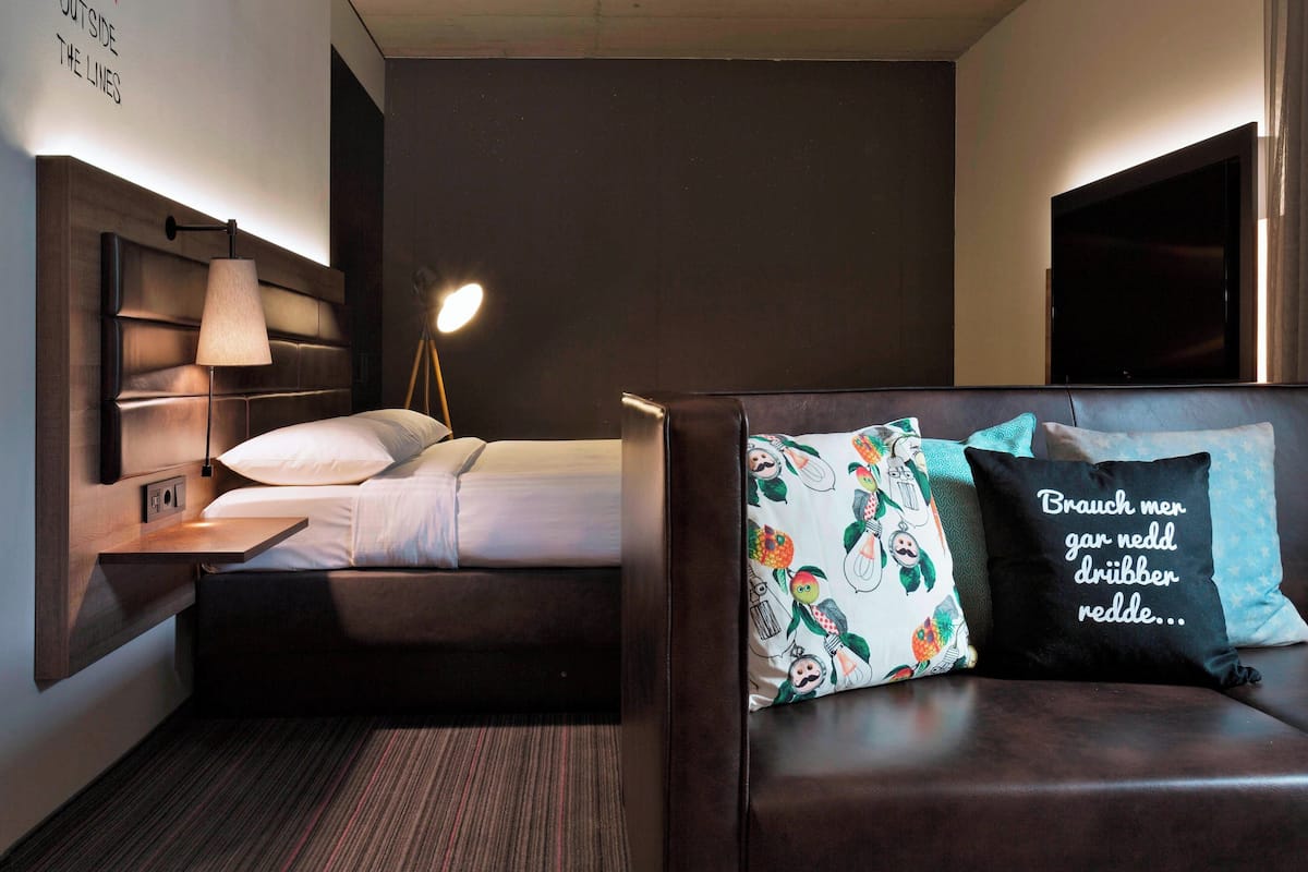 Una habitación de hotel moderna con cama, lámpara de noche y sofá con cojines decorativos.