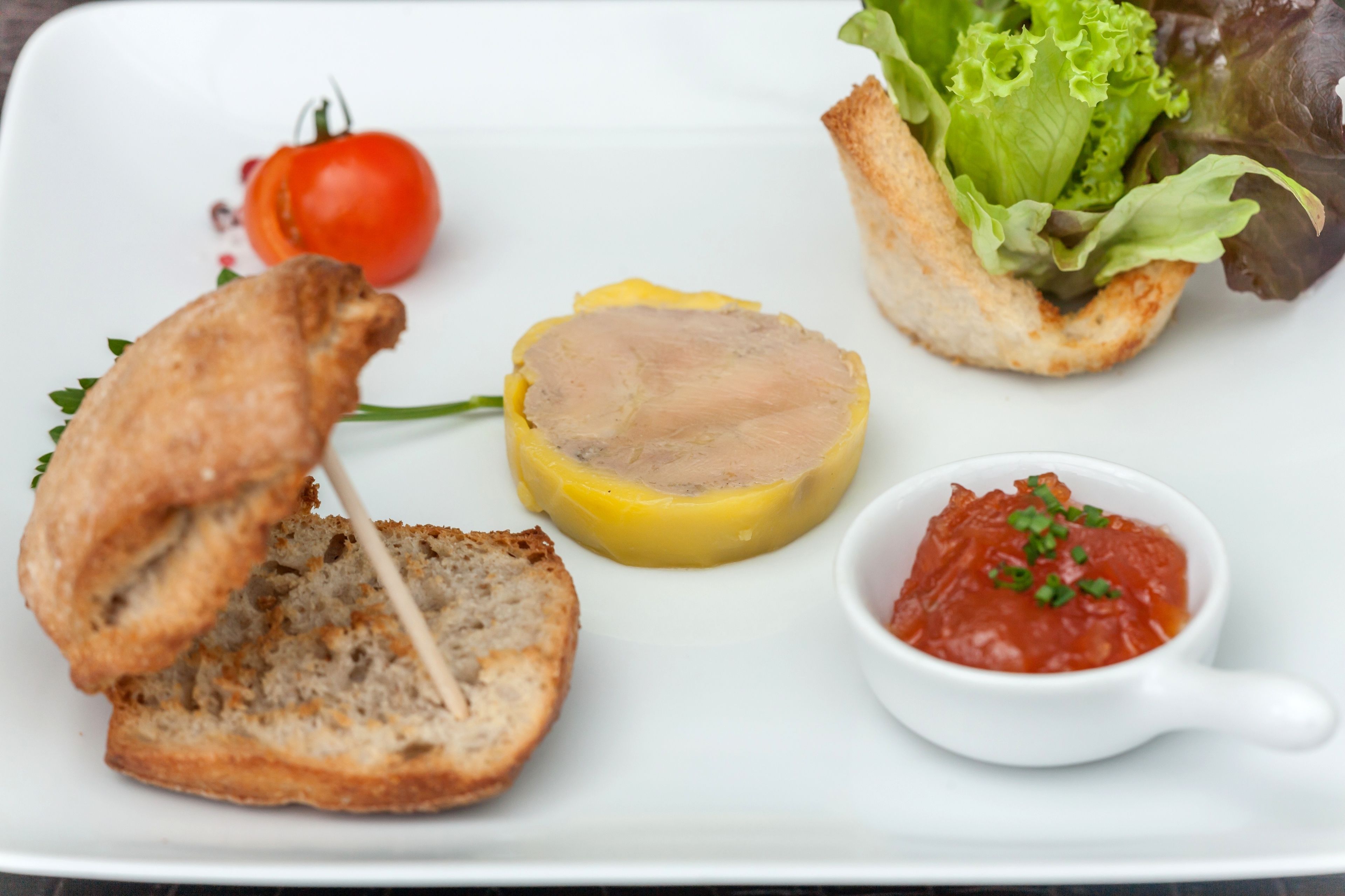 A plate with a slice of bread, a round meat slice, a cherry tomato, a lettuce leaf, and a small bowl of sauce.