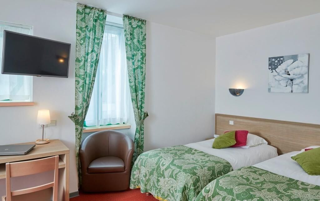 Ein Hotelzimmer mit Schreibtisch, Stuhl, zwei Betten, Fernseher und einem Vorhangfenster.