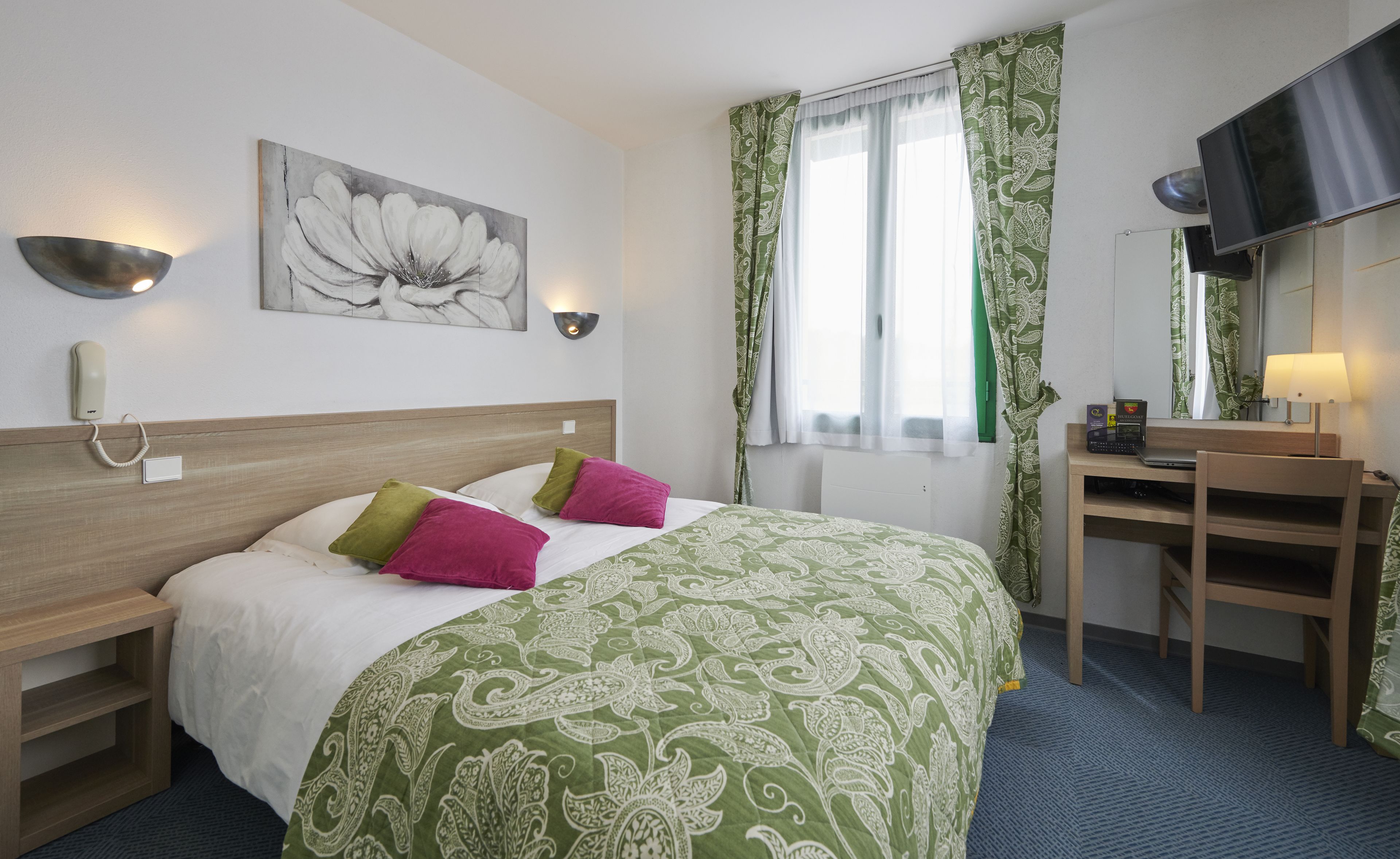 Una camera d'albergo con un letto, una scrivania, una sedia, un telefono, un quadro e una finestra con tende.