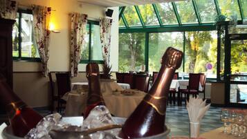 A restaurant interior with tables set for dining, a large window with a view of trees, and a bucket with champagne bottles and ice.