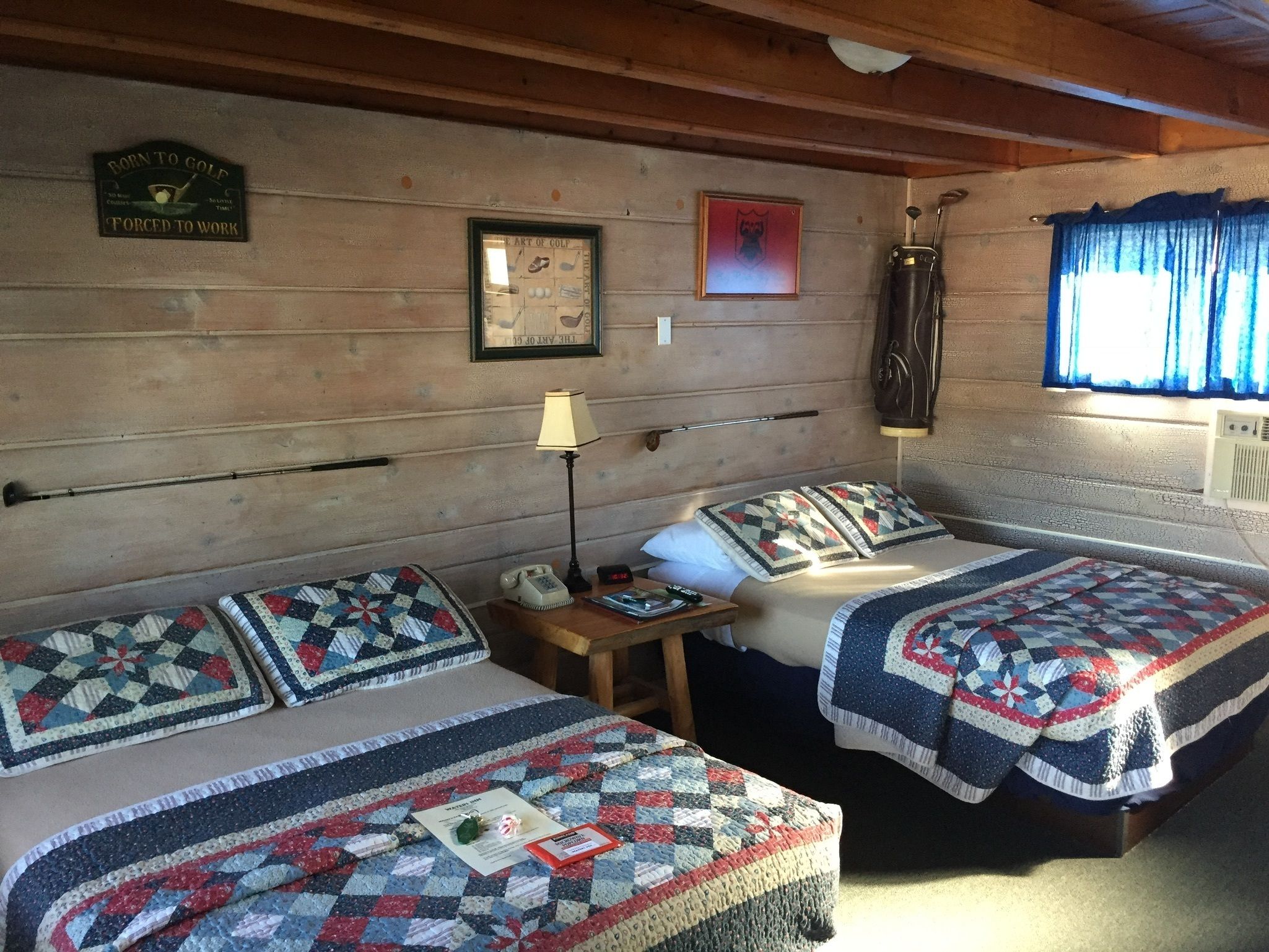 A room with two beds, each with a quilt, a lamp, a telephone, and a framed picture on the wall.