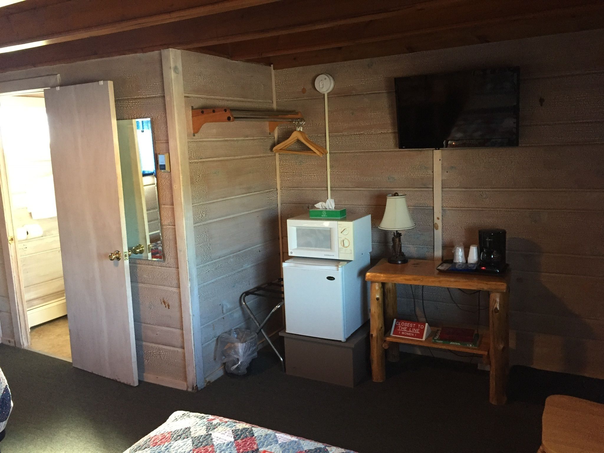 A small, cozy room with a wooden interior, a microwave, a small table with a lamp, and a television mounted on the wall.