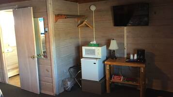 A small, cozy room with a wooden interior, a microwave, a small table with a lamp, and a television mounted on the wall.