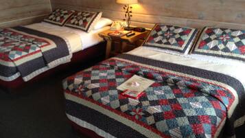 Ein Hotelzimmer mit zwei Betten, jedes mit einer gesteppten Tagesdecke und Kissen. Dazwischen befindet sich ein Nachttisch mit Lampe und einer Uhr.