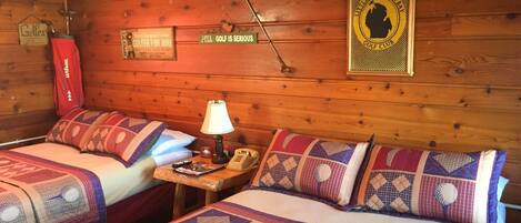 A cabin-style room with two beds, a lamp, a telephone, and a framed sign on the wall.