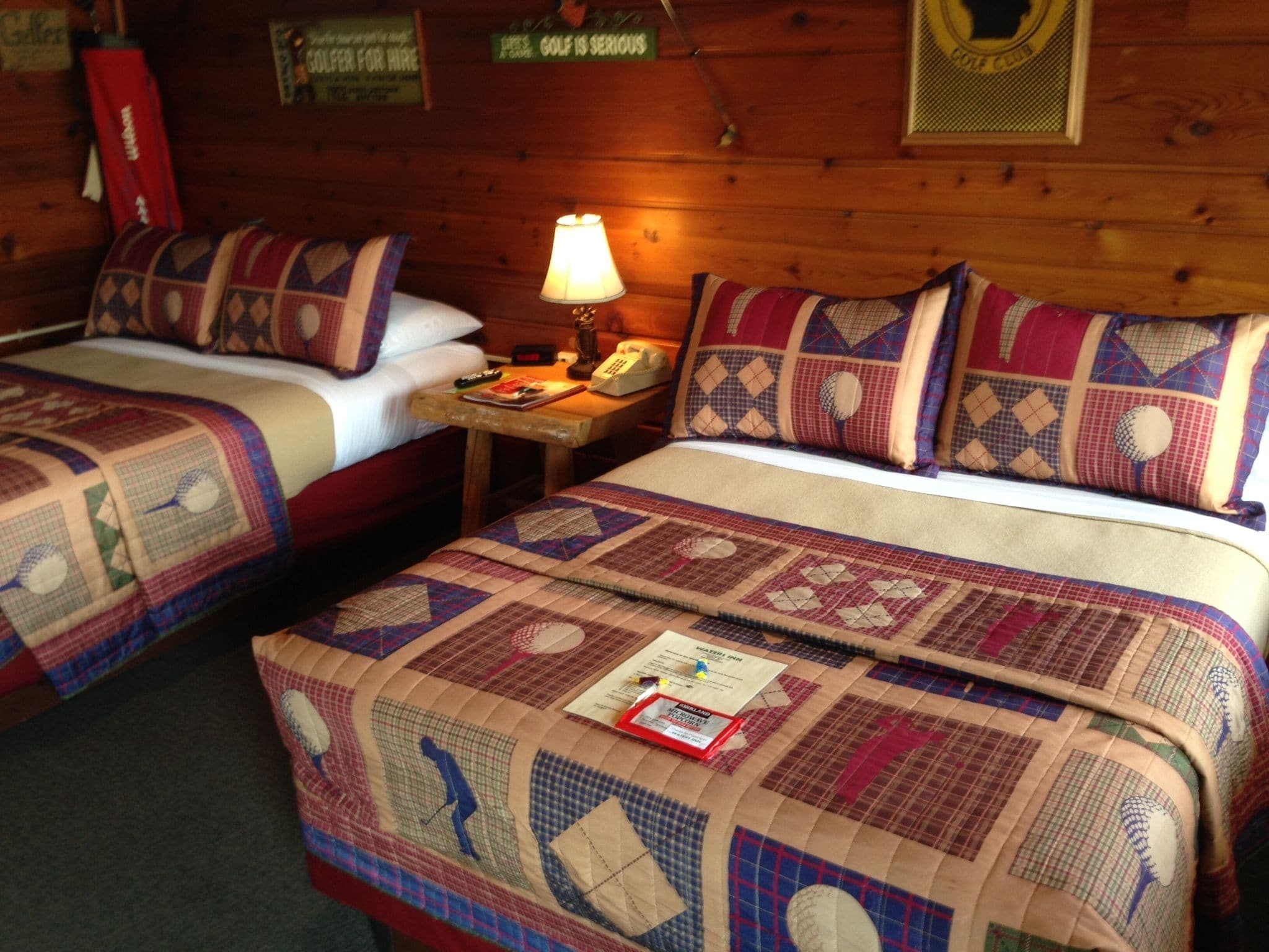 A hotel room with two beds, each with a quilted bedspread and pillows. There is a nightstand with a lamp, a telephone, and a book. The room has wooden walls and a sign that says "GOLF CLUB".